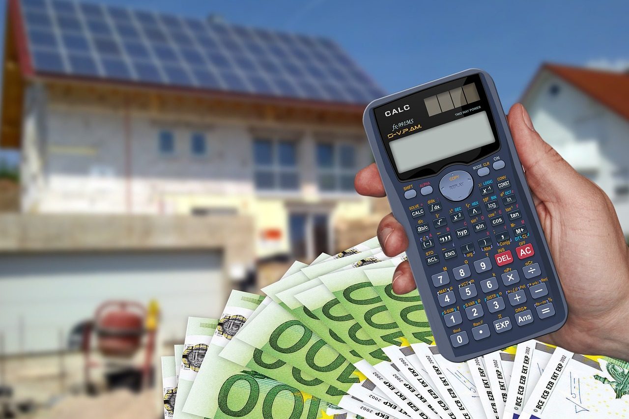 Das Bild zeigt eine Hand, die einen Taschenrechner hält, während im Hintergrund ein Haus mit installierten Solarmodulen zu sehen ist. Vor dem Taschenrechner sind mehrere 100-Euro-Scheine deutlich sichtbar. Das Bild symbolisiert die finanzielle Planung und Kalkulation, die mit der Installation einer Photovoltaikanlage verbunden sind. Es stellt die Frage „Was kostet eine 10 kWp PV-Anlage mit Speicher?“ in den Vordergrund, indem es die Verbindung zwischen den Kosten, den möglichen Investitionen und den potenziellen Einsparungen darstellt, die eine solche Anlage mit sich bringen kann. Das Haus im Hintergrund verdeutlicht, dass es sich um eine praktische Anwendung dieser Technologie auf Einfamilienhäusern handelt, während der Taschenrechner und das Geld die wirtschaftlichen Überlegungen betonen, die bei der Entscheidung für eine Solaranlage berücksichtigt werden müssen.