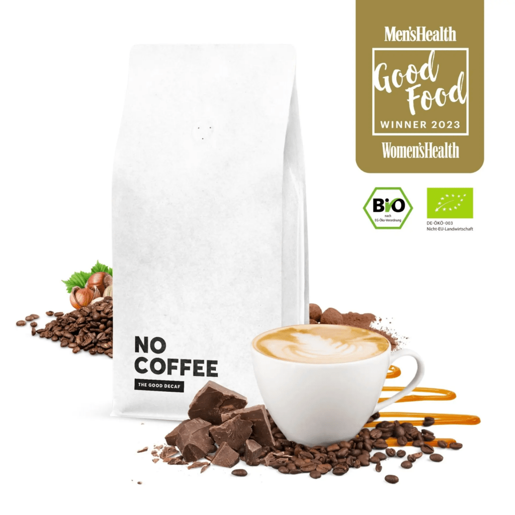 Das Bild zeigt eine minimalistisch gestaltete Szene, die den koffeinfreien Kaffee von "No Coffee" präsentiert. Im Mittelpunkt steht eine weiße Kaffeeverpackung mit dem Aufdruck "NO COFFEE" und dem Zusatz "THE GOOD DECAF". Um die Verpackung herum sind Kaffeebohnen, Schokoladenstücke und eine Tasse Cappuccino arrangiert, um die geschmacklichen Noten des Kaffees zu betonen.

Links neben der Verpackung liegen Kaffeebohnen und einige Haselnüsse, während sich rechts unten Schokoladenstücke und eine Tasse Cappuccino befinden, umgeben von Kaffeebohnen.

Auf der rechten Seite des Bildes befinden sich mehrere Logos, die Auszeichnungen und Zertifizierungen darstellen. Dazu gehört das "Men’s Health Good Food Winner 2023" und das "Women’s Health Good Food Winner 2023" Siegel, das auf die Auszeichnung des Produkts hinweist. Darunter sind Bio-Zertifizierungen zu sehen, die die ökologische Herstellung des Kaffees bestätigen.

Die gesamte Szene ist in hellen, einladenden Farben gehalten, die eine hochwertige und saubere Ästhetik vermitteln.