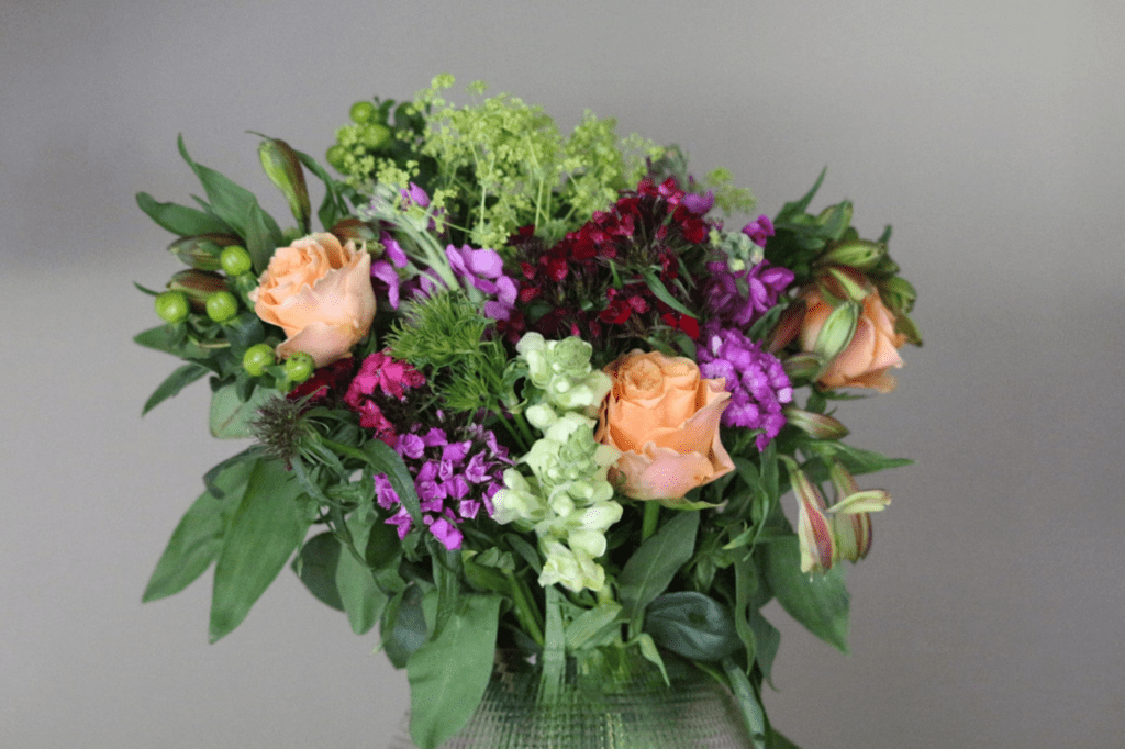 Auf dem Bild ist ein farbenfroher Blumenstrauß zu sehen, der aus verschiedenen Blumenarten und Pflanzen besteht. Der Strauß enthält zart orangefarbene Rosen, die sanft zwischen den kräftigeren Farben hervorstechen. Daneben finden sich dunkelrote Blüten, violette und weiße Blumen, die dem Arrangement Tiefe und Lebendigkeit verleihen. Grünblättrige Zweige und zarte Gräser ergänzen den Strauß und geben ihm eine natürliche, frische Optik. Insgesamt wirkt das Arrangement lebendig, harmonisch und gleichzeitig elegant – perfekt für einen festlichen Anlass.